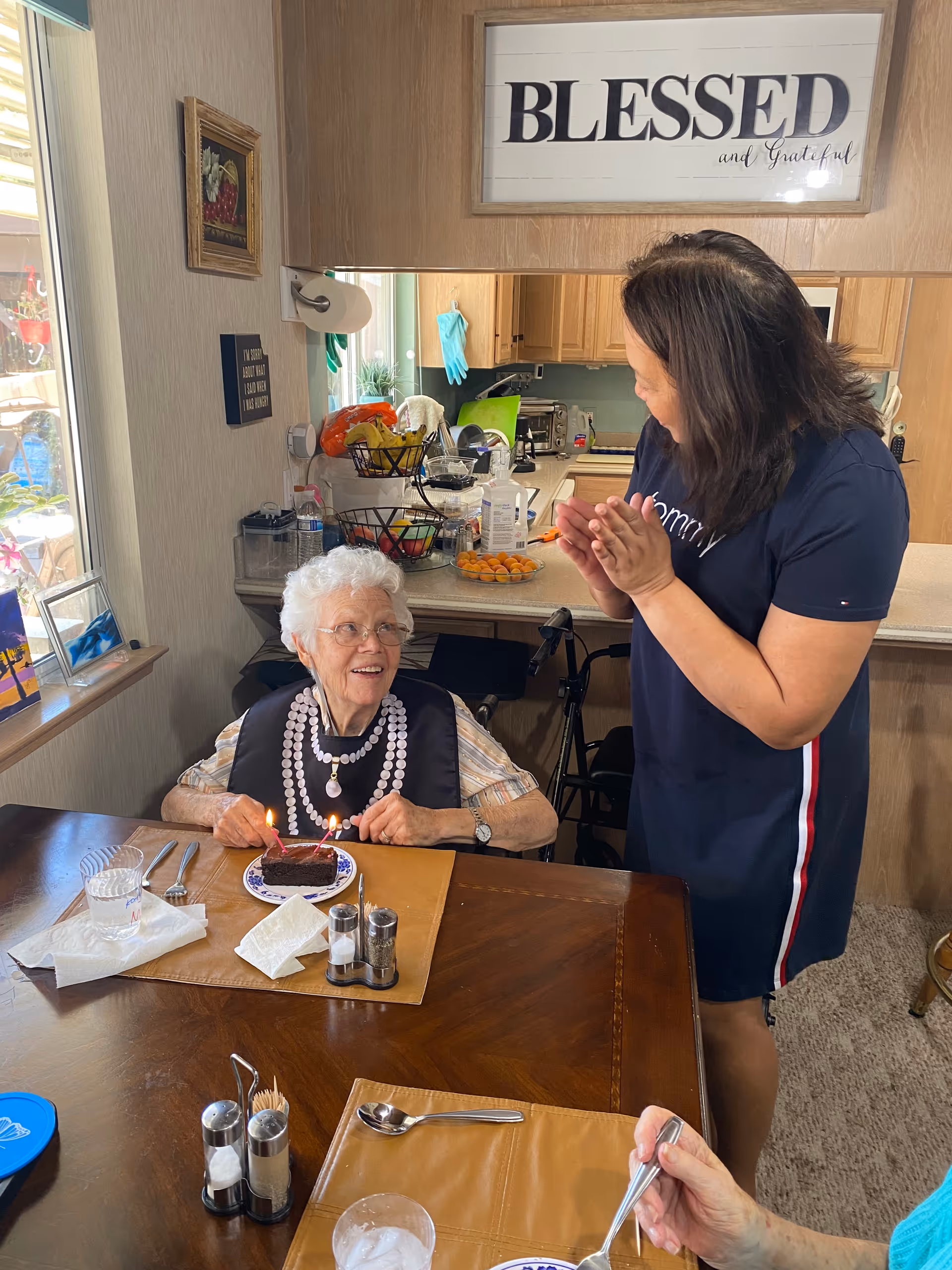 An elderly woman with white hair and glasses sits at a dining table with a small chocolate cake with lit candles in front of her. A younger woman stands nearby, clapping and smiling at the elderly woman. The setting appears to be a cozy kitchen with wooden cabinets and various kitchen items on the counter. A framed sign above the kitchen reads 'BLESSED and Grateful.'