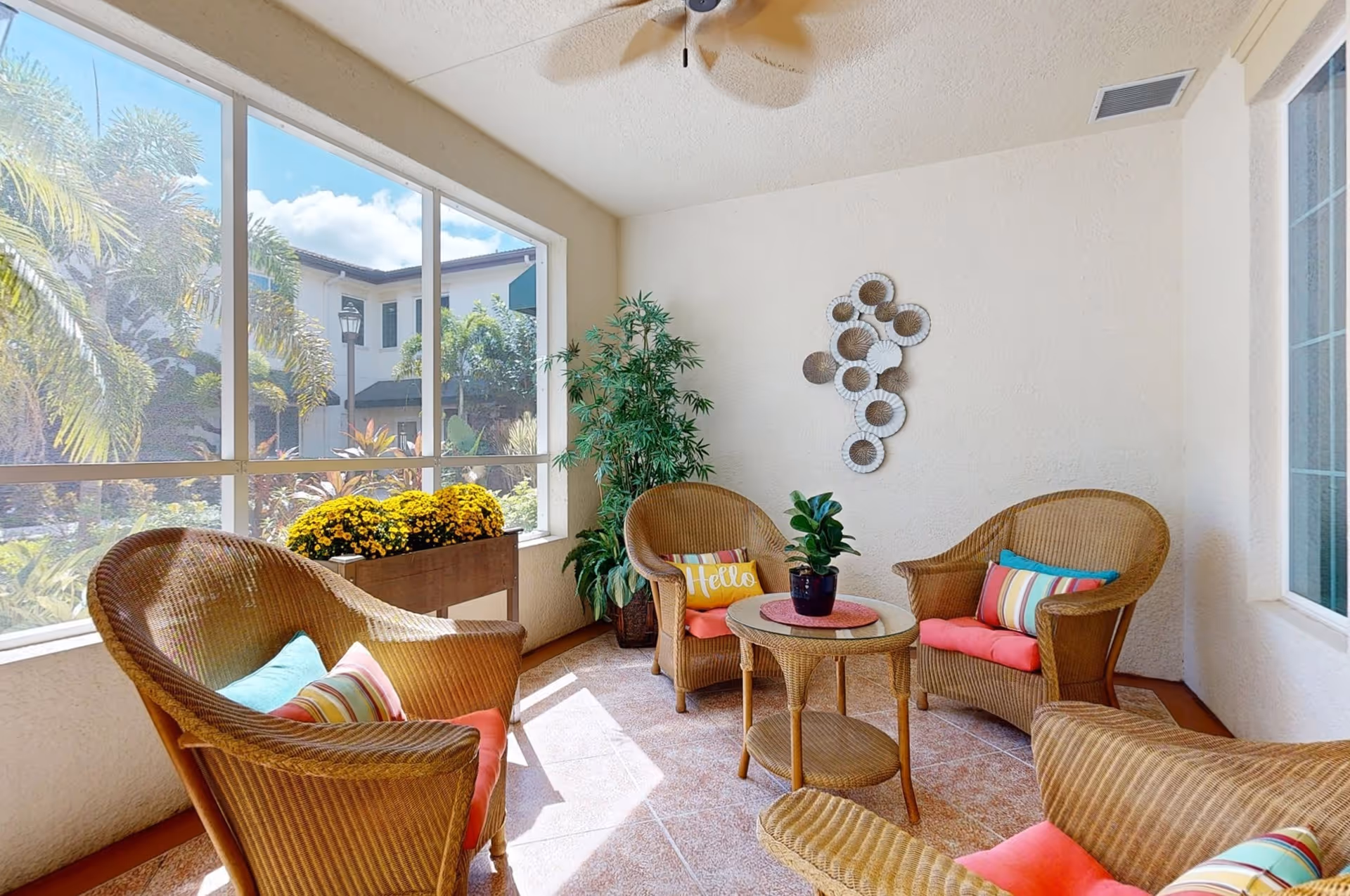 A bright and cozy indoor seating area with four wicker chairs featuring colorful cushions and pillows, a small round wicker table with a potted plant, a decorative wall hanging, and large windows showing an outdoor garden with palm trees and a building in the background.