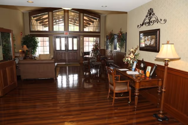 Interior view of a nursing and rehabilitation center lobby with polished wooden floors, a wooden desk with a chair and lamp, floral decorations, a seating area with sofas, and large glass double doors letting in natural light.