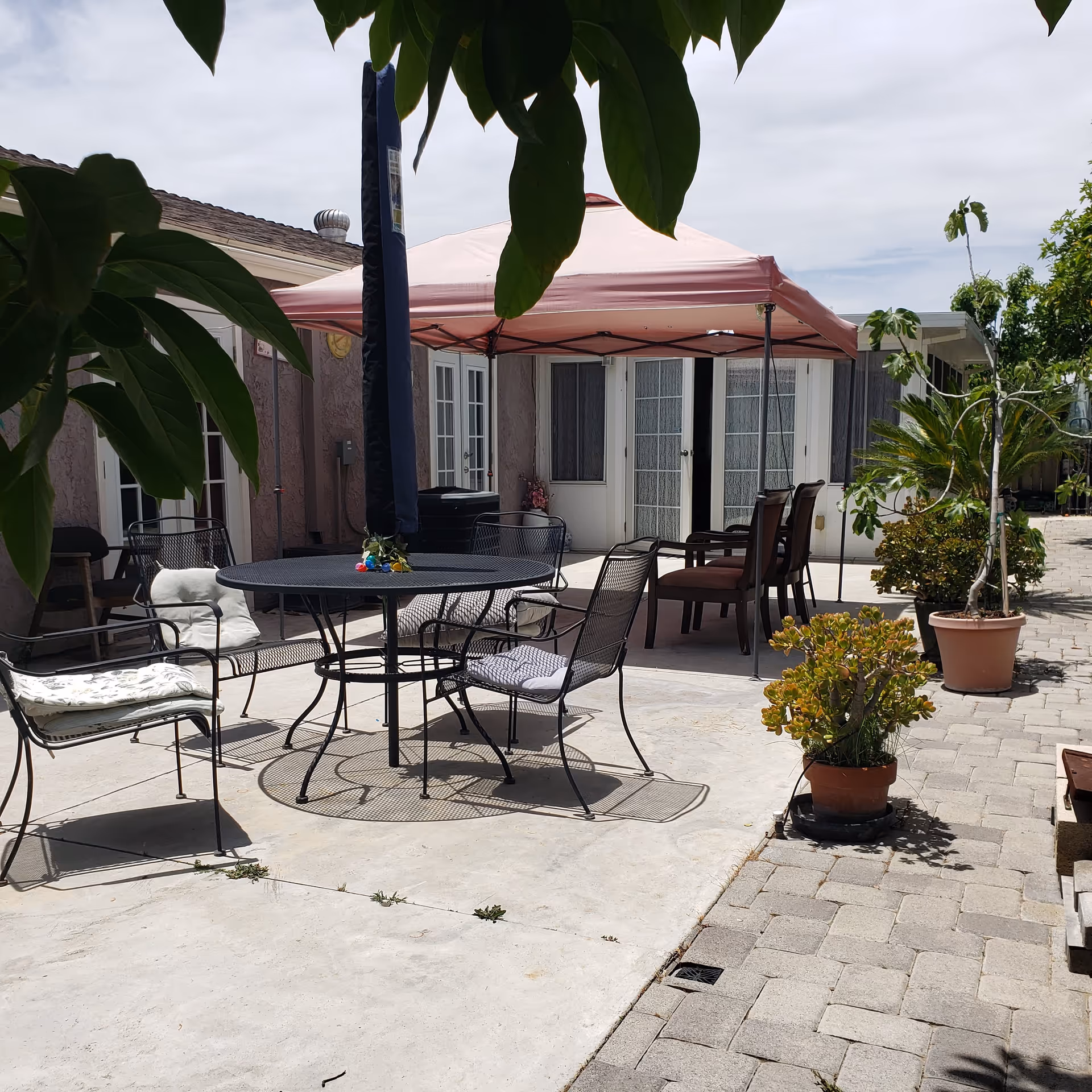 Outdoor patio area with a round metal table and four metal chairs with cushions. A pink canopy tent provides shade over two wooden chairs and a small table. Potted plants and trees surround the paved patio area, with a building featuring multiple doors and windows in the background. Green leaves partially frame the top of the image.