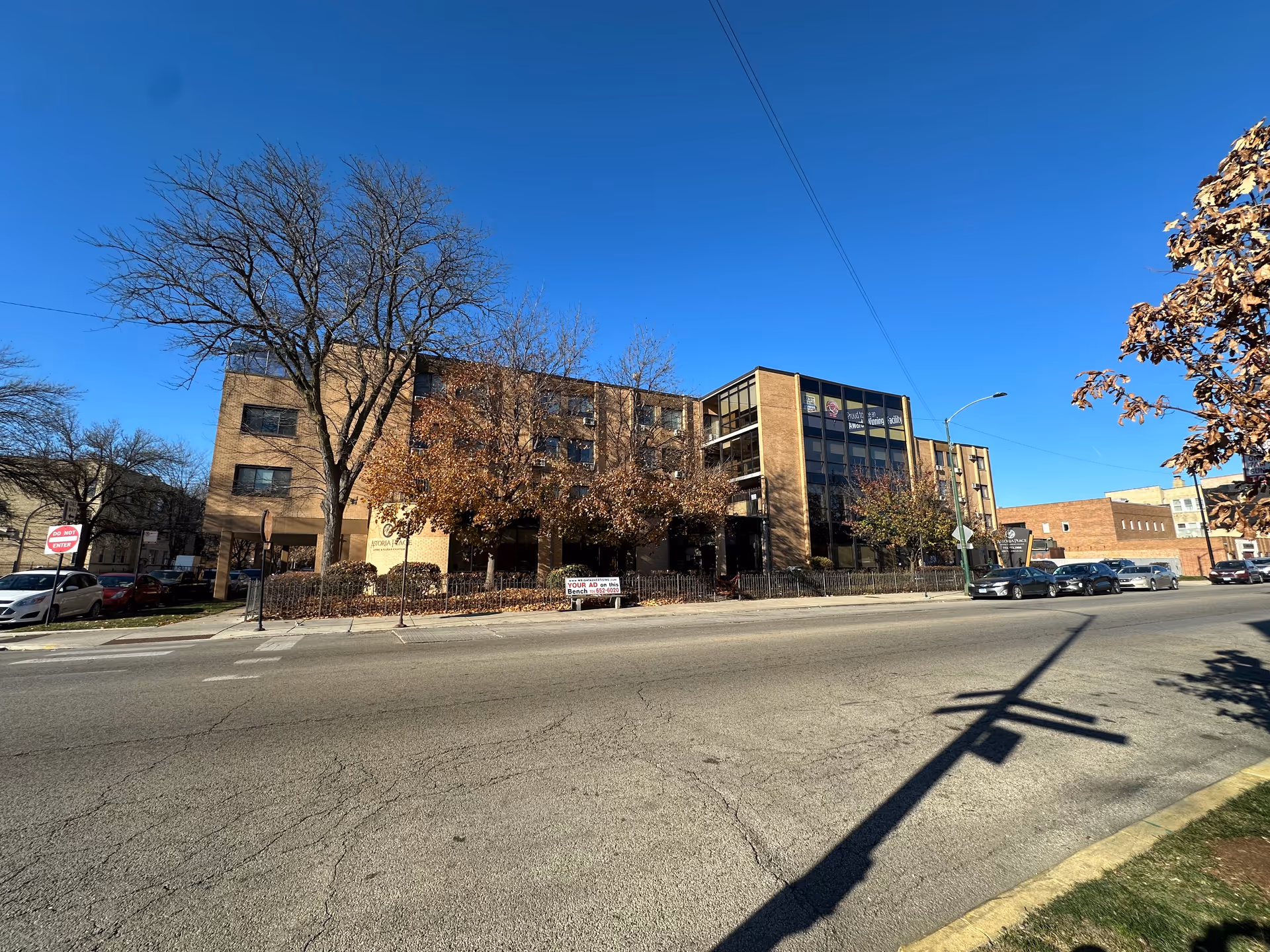 Exterior view of a multi-story brick building with large windows, surrounded by leafless and autumn-colored trees, parked cars along the street, and a clear blue sky overhead.
