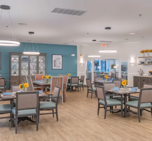 A bright and spacious dining room in a senior living facility with multiple tables and chairs arranged neatly. Each table is set with blue napkins and small vases containing yellow flowers. The room features light wood flooring, teal and white walls, modern hanging light fixtures, and a glass-front cabinet against one wall. Large windows and glass doors allow natural light to enter the space.