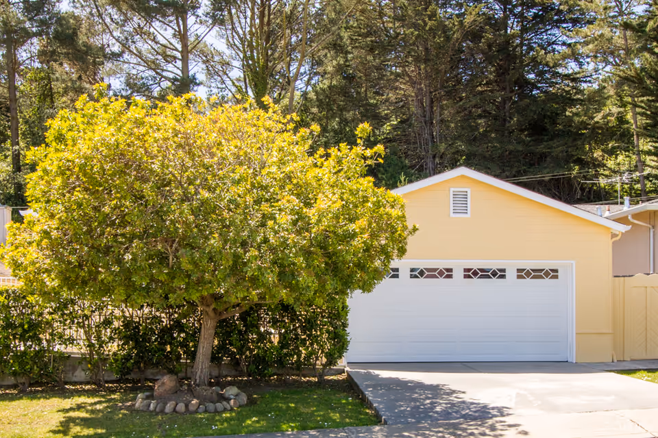 A yellow garage with a white door featuring decorative windows, situated next to a green leafy tree surrounded by a small circular arrangement of rocks, with a background of tall trees and a clear sky.