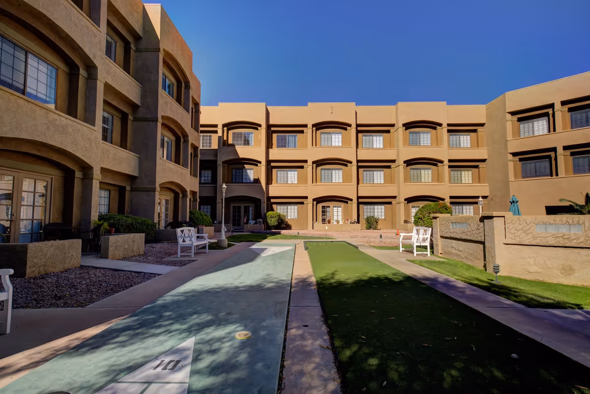 Outdoor courtyard area of a senior living facility with a shuffleboard court in the center, surrounded by beige multi-story buildings with multiple windows. There are white benches and some greenery along the sides under a clear blue sky.