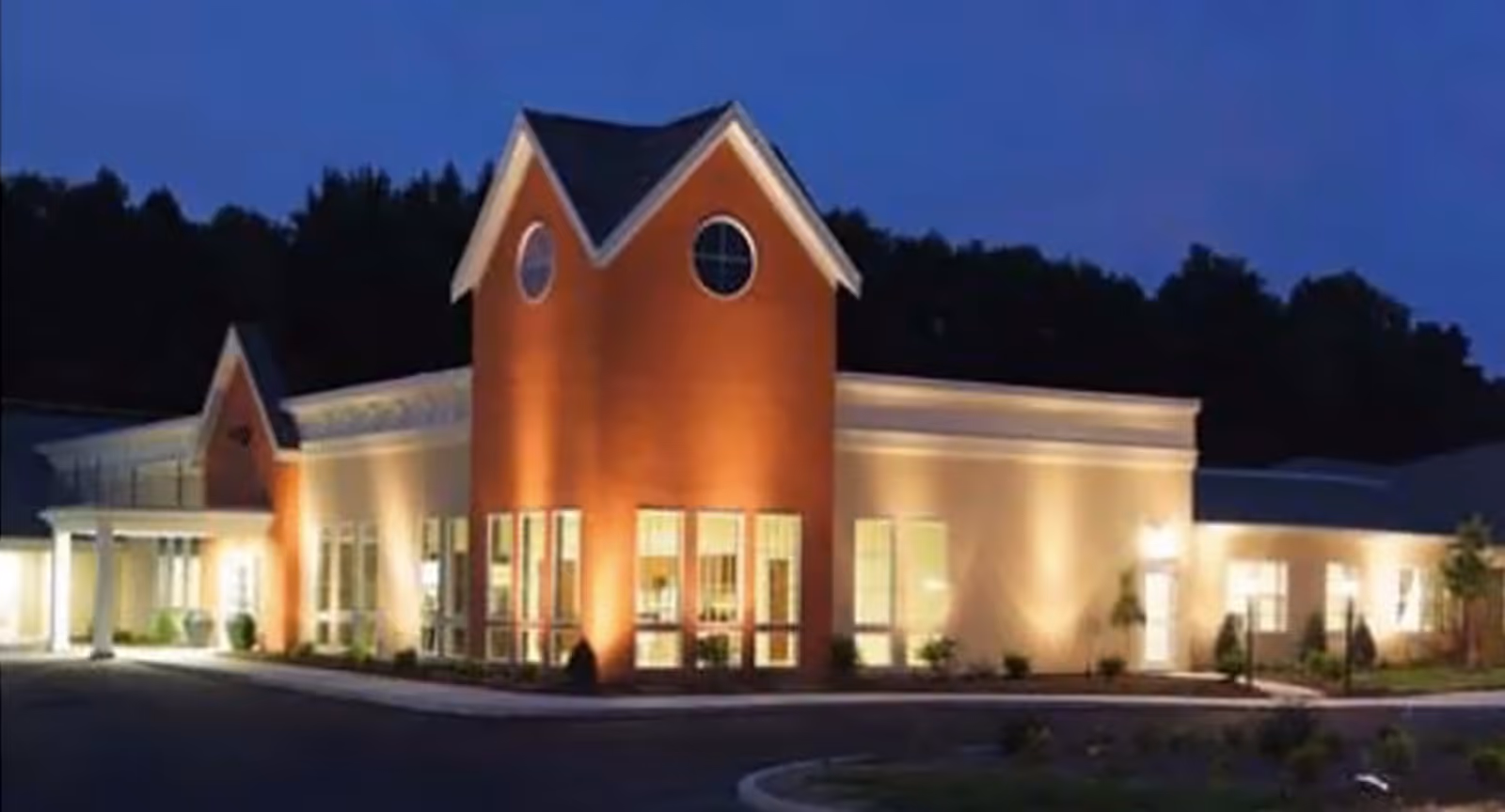 Exterior view of a well-lit nursing center building at dusk with a prominent brick tower featuring two round windows and large glass windows along the front.