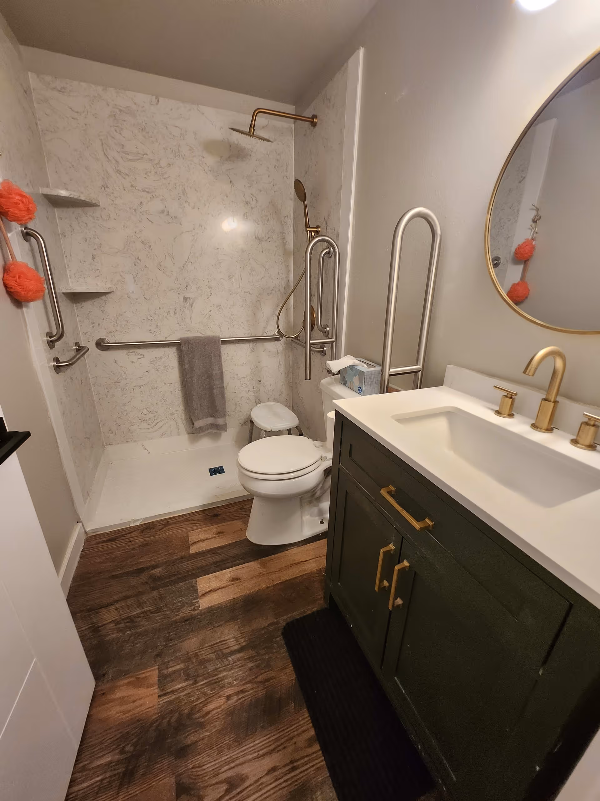 A bathroom with a walk-in shower featuring a rain showerhead and handheld shower, grab bars, a toilet with a safety frame, a green vanity with a white countertop and gold faucet, a round mirror, and wooden flooring.