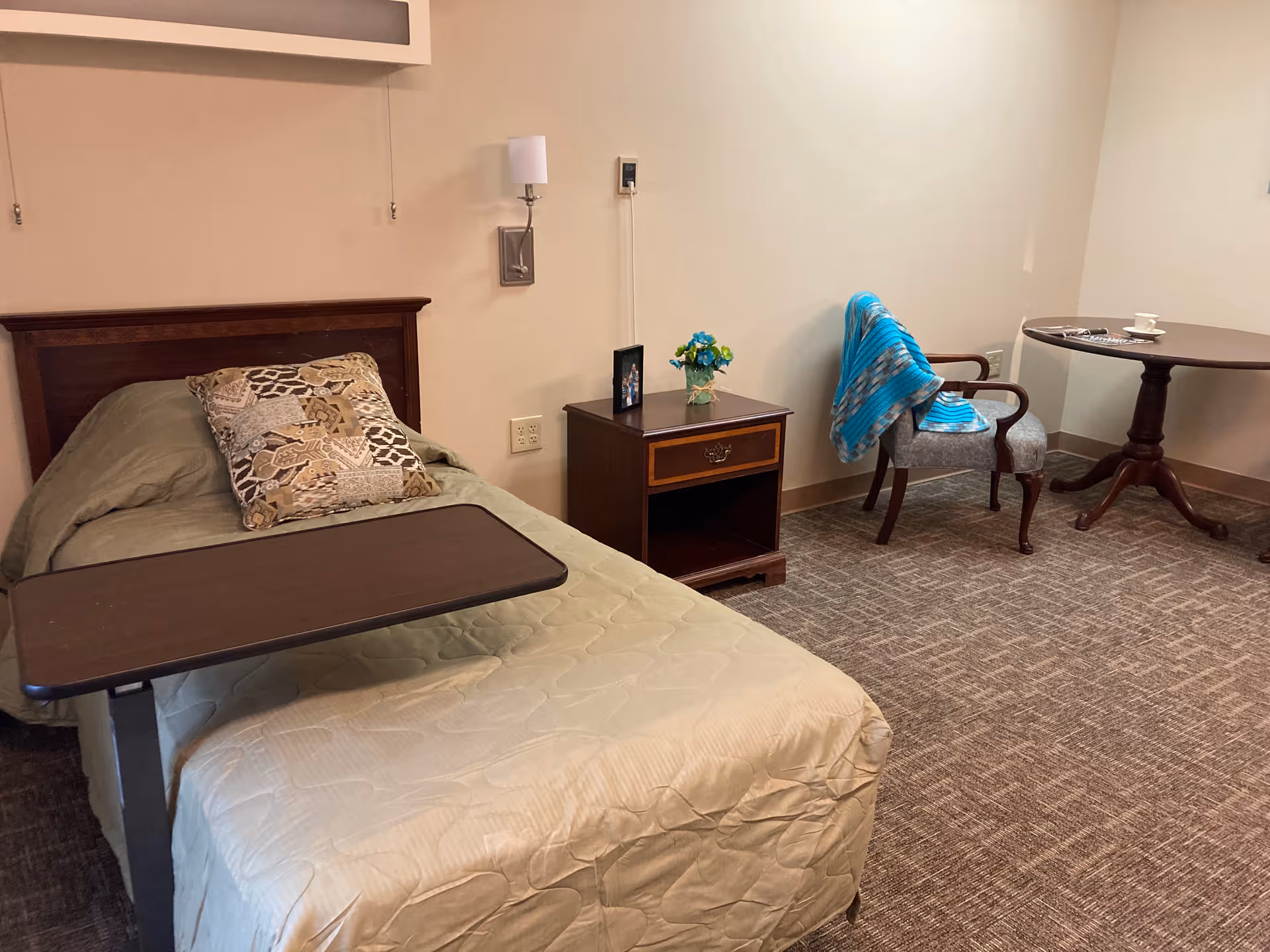 A single bed with a beige quilt and a patterned pillow, a dark wooden overbed table, a wooden nightstand with a small flower vase and a framed photo, a gray upholstered chair with a blue striped blanket draped over it, and a round wooden table with a cup and saucer on top, all set on a carpeted floor in a softly lit room.