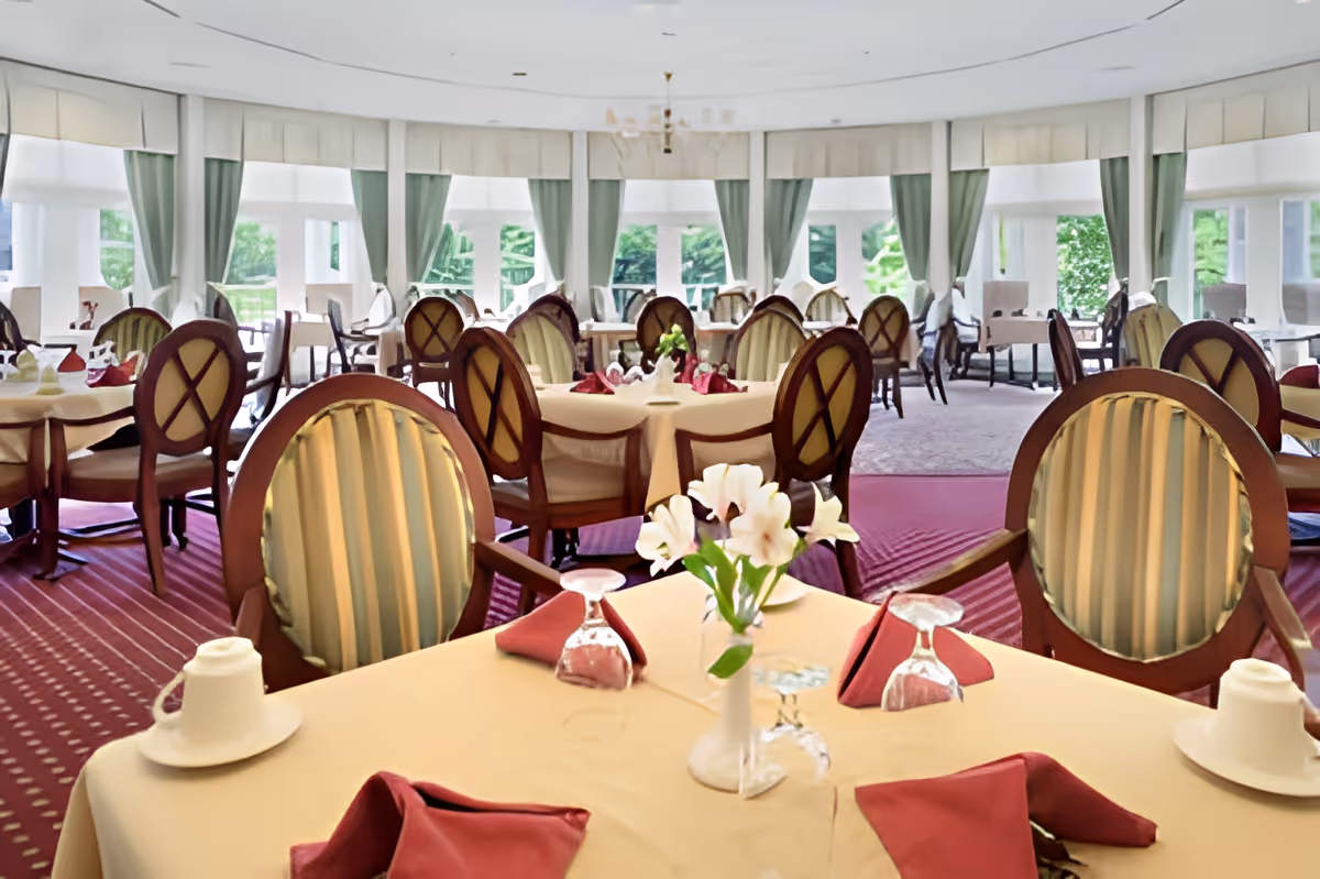 A spacious dining room with round tables covered in beige tablecloths, each set with upside-down wine glasses, folded red napkins, and white flower centerpieces. The room features elegant wooden chairs with striped upholstery, large windows with green curtains, and a red carpeted floor.