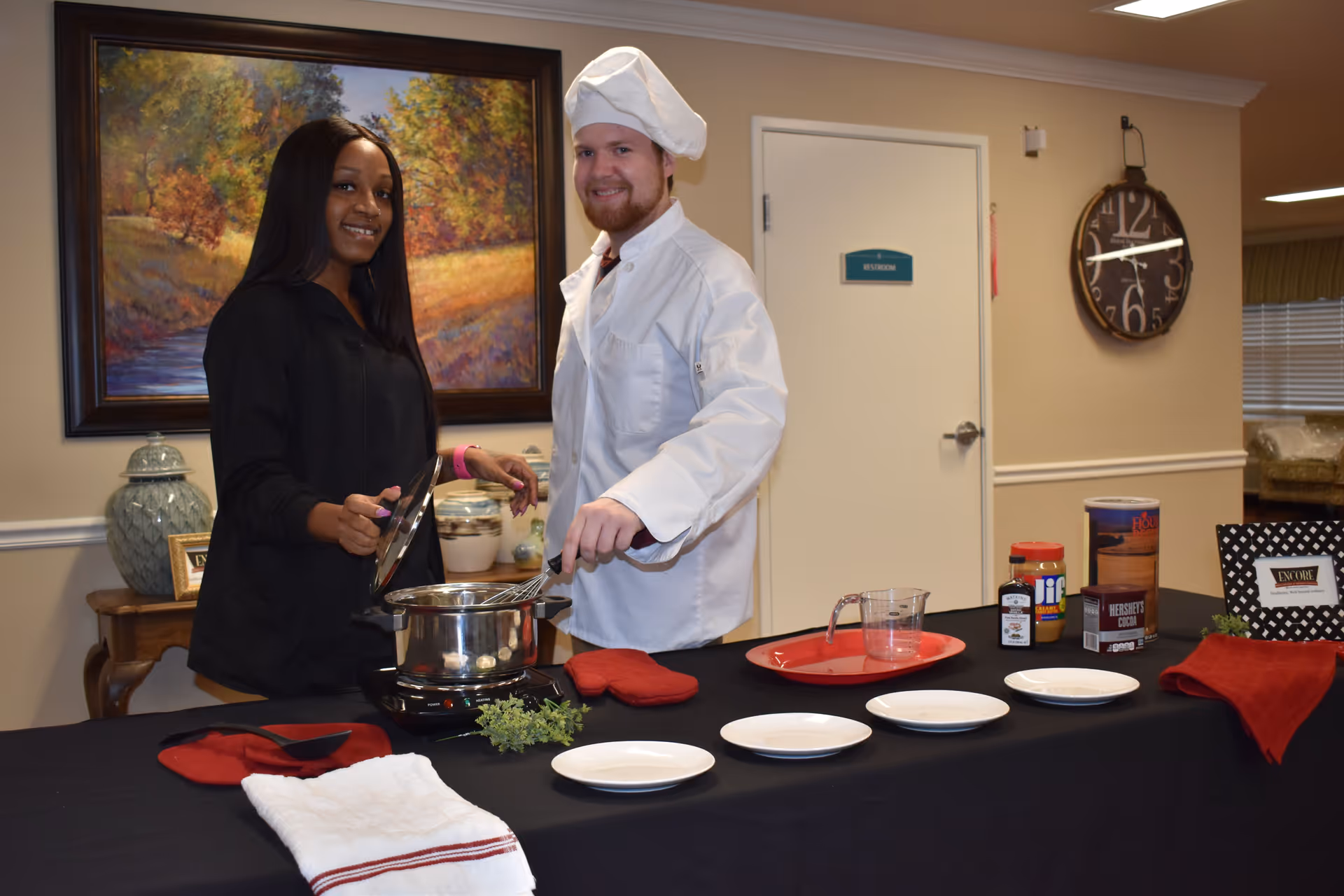 A woman and a man dressed as a chef stand behind a table with cooking supplies including a pot on a portable stove, plates, and ingredients like peanut butter and cocoa. The setting appears to be an indoor room with a painting on the wall and a clock showing the time as 12:15.