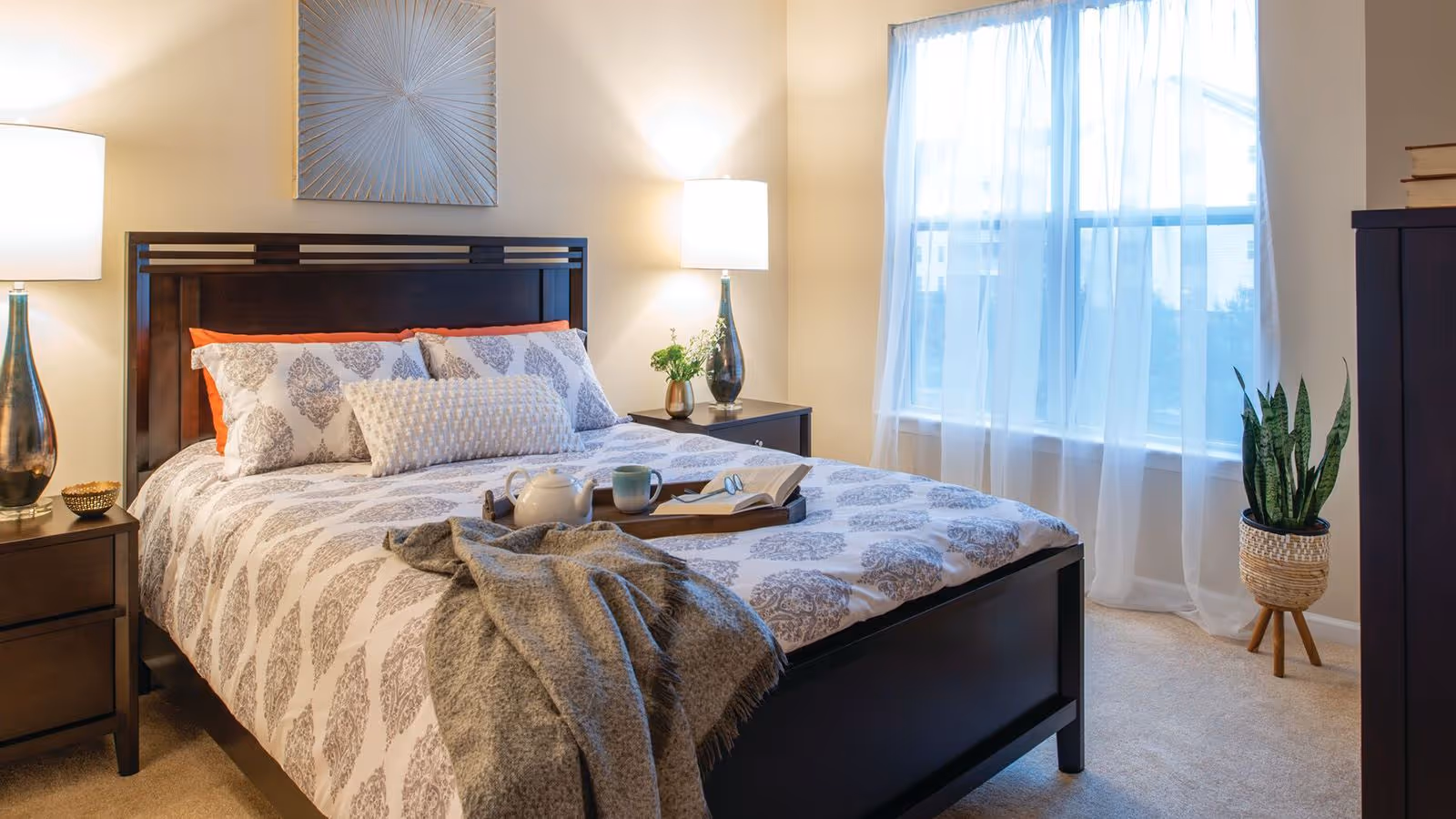 A cozy bedroom with a dark wooden bed frame and matching nightstands on each side. The bed is made with patterned white and gray bedding, accented by orange pillows and a textured gray throw blanket. A tray with a teapot, cup, and an open book rests on the bed. Two table lamps with white shades provide warm lighting. A large window with sheer white curtains lets in natural light. A potted plant stands near the window, and a decorative wall art piece hangs above the headboard.