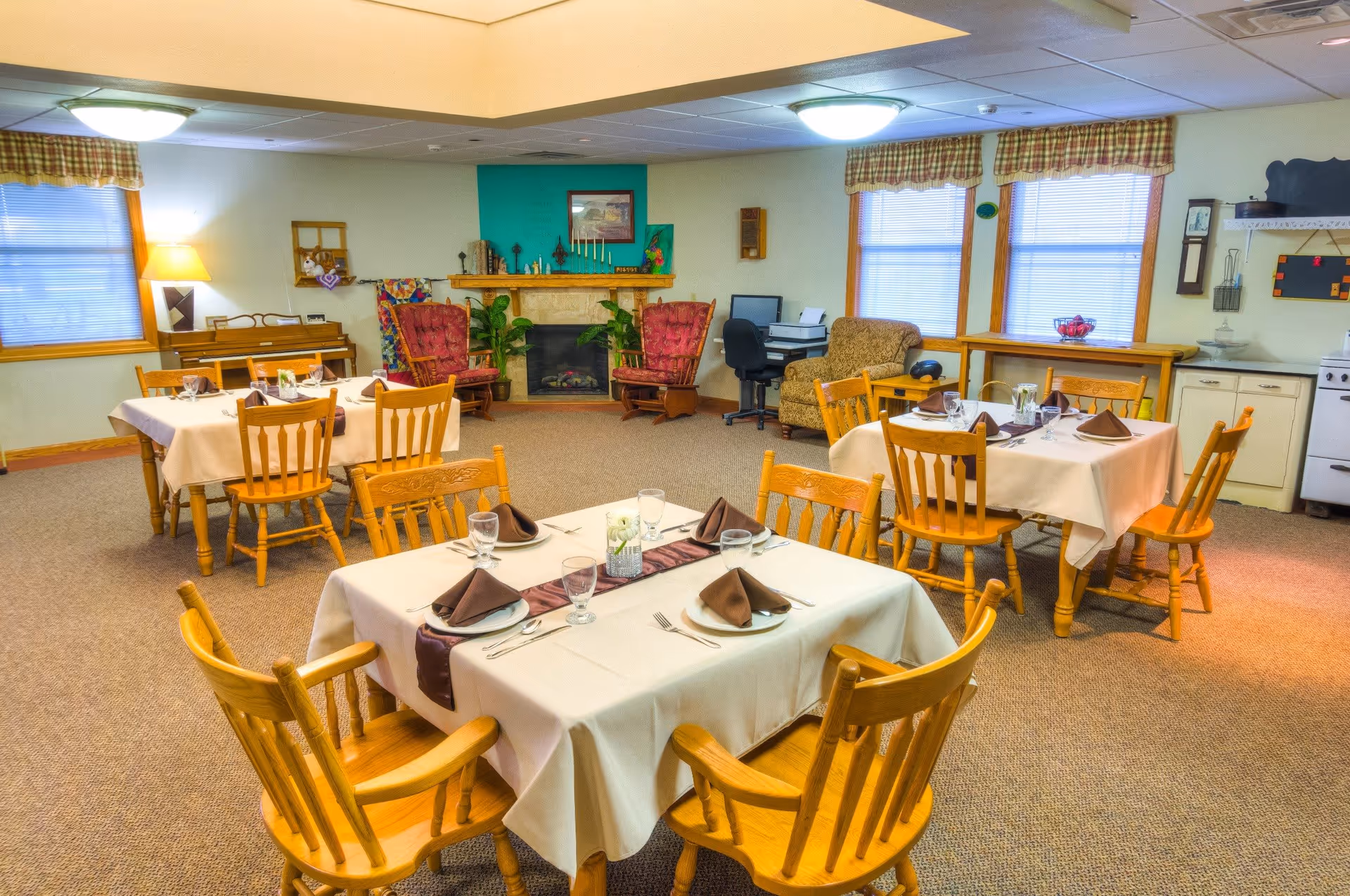 A cozy dining room in a senior living facility with four wooden tables covered with white tablecloths, each set with plates, glasses, and brown napkins. The room features wooden chairs, a fireplace with two red cushioned rocking chairs, a piano, a computer desk with a chair, and windows with plaid valances letting in natural light.