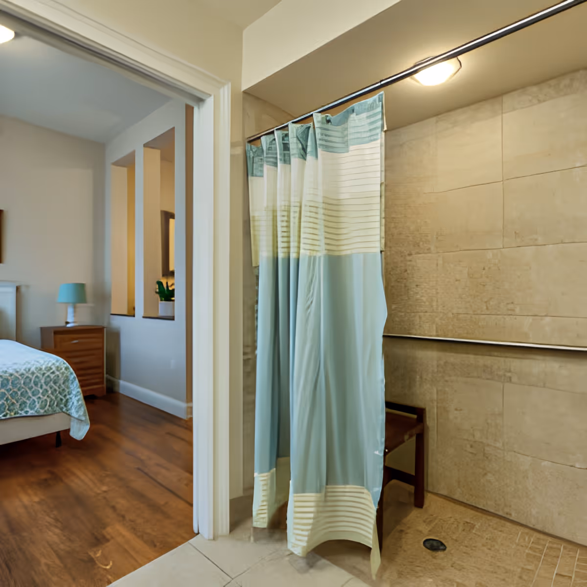 View of a bathroom shower area with a blue and white striped shower curtain partially drawn, beige tiled walls, and a wooden bench inside the shower. Adjacent to the bathroom is a bedroom with wooden flooring, a bed with a patterned bedspread, a nightstand, and a lamp.