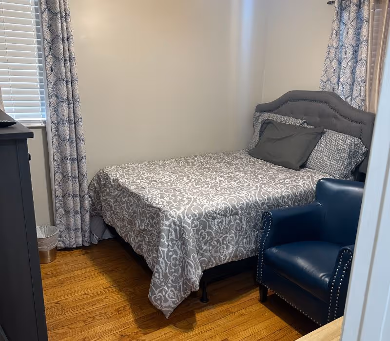 Small bedroom with patterned bedspread, blue chair, dresser, curtains, and hardwood floor.