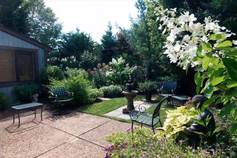 A peaceful outdoor garden area with metal chairs and a small table on a stone patio surrounded by lush green plants, flowering bushes, and trees. A stone birdbath or fountain is centered on a small paved square in the garden.