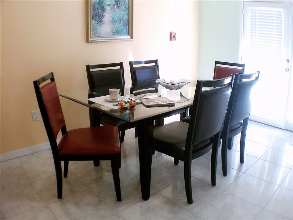 Glass-top dining table set with six chairs, a centerpiece and a cup in a bright tiled dining area with a door to the outside.