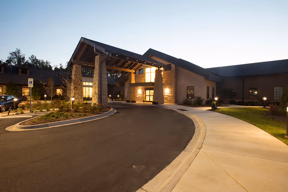 Front entrance of a lit health care building with a covered porte-cochère, curved driveway and landscaped walkway at dusk.