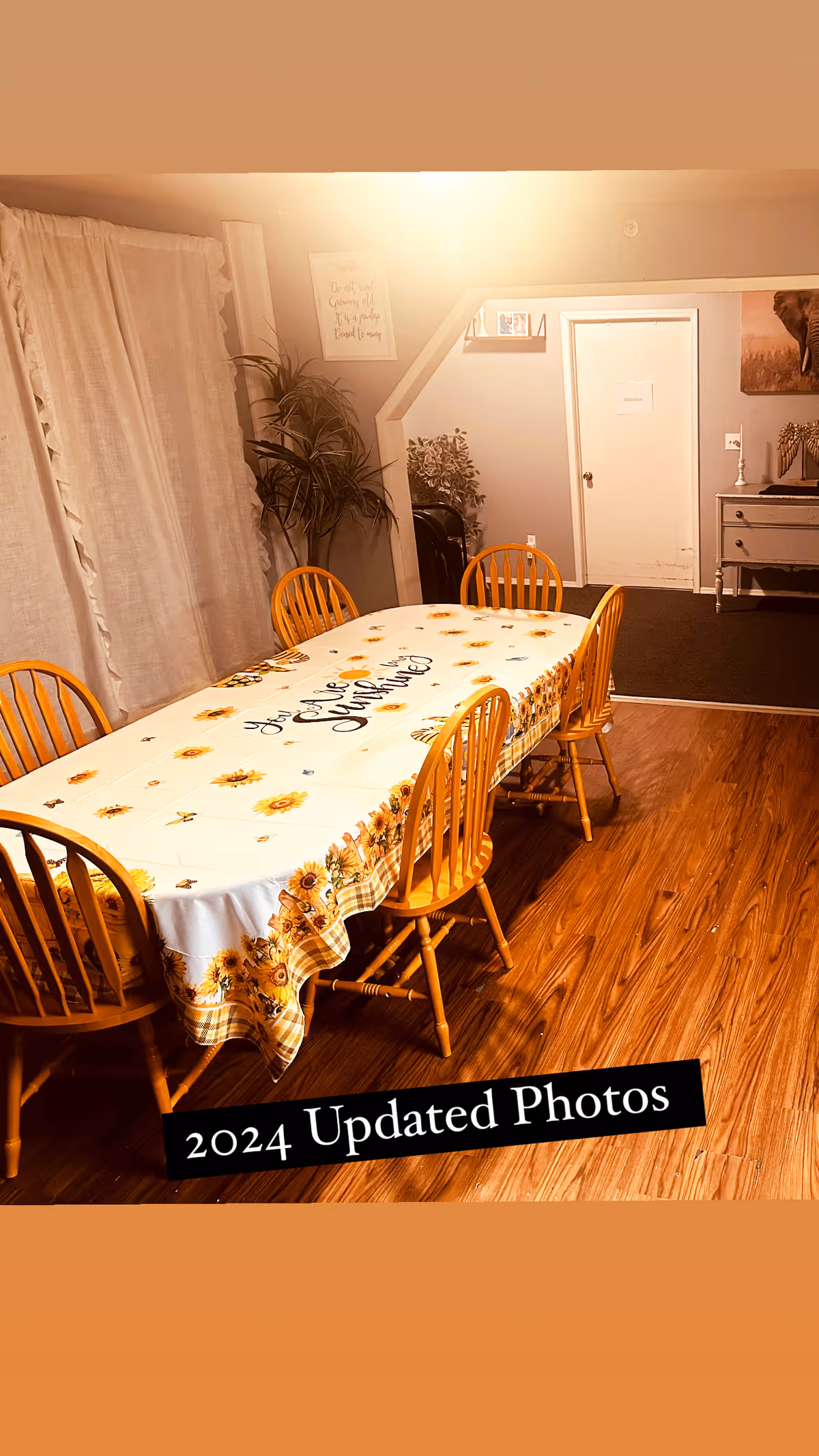 A dining room with a wooden table covered by a sunflower-themed tablecloth that reads 'You are my sunshine'. Six wooden chairs surround the table. The room has wooden flooring, a white curtain on the left, a potted plant in the corner, and a white door and dresser in the background.