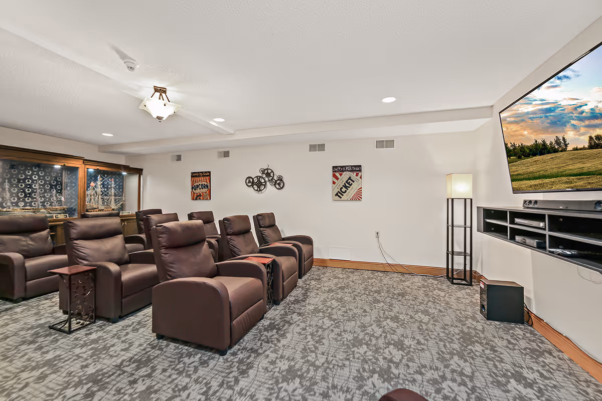 A cozy home theater room with multiple brown leather recliner chairs arranged in rows facing a large flat-screen TV mounted on the wall. The room has patterned carpet, a floor lamp in the corner, and decorative wall art including a popcorn sign and a ticket poster. A wooden cabinet with glass doors is visible in the back corner.