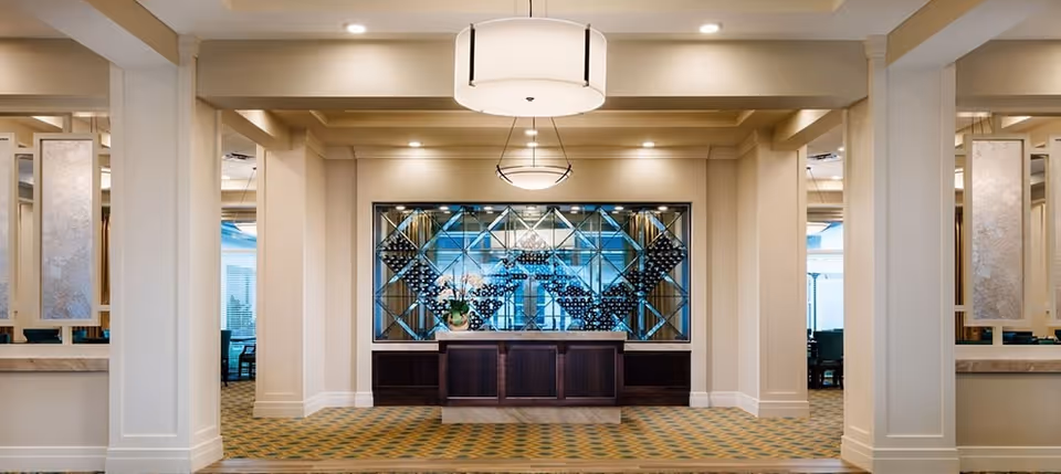 Interior view of a senior living facility lobby or common area with a central wooden reception desk in front of a decorative mirrored wall featuring a geometric pattern. The space is well-lit with ceiling lights and has beige walls, columns, and patterned carpet flooring. Seating areas are visible through openings on both sides.