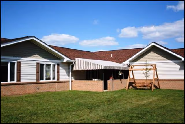 Exterior view of a single-story building with light-colored siding and a brick base. The building has a striped awning over a patio area and a wooden swing set on a grassy lawn under a blue sky with some clouds.