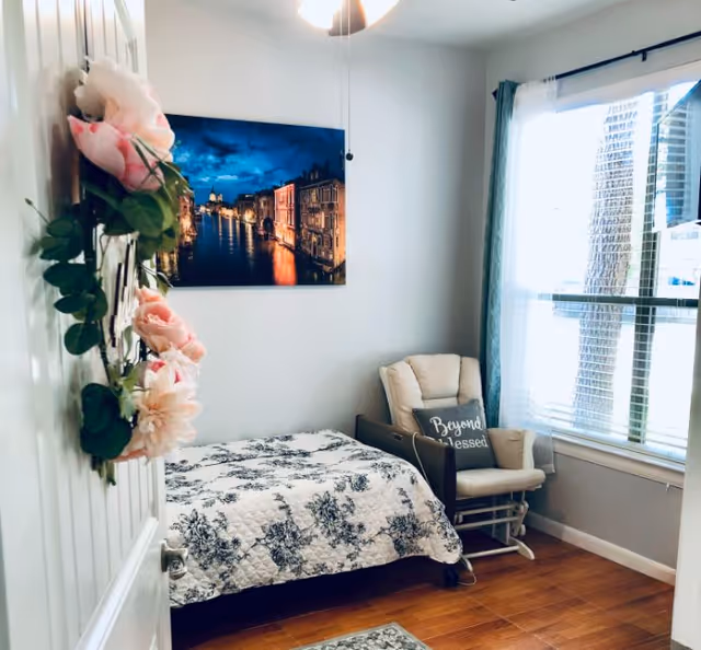 Small, bright bedroom with a single bed covered in a floral quilt, a padded glider chair by a window, wall art, and a floral wreath on the door.