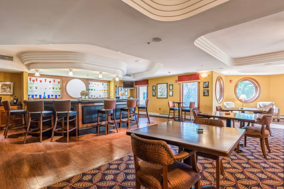 Bright senior living common room with a curved bar, high stools, dining tables and chairs, and round windows letting in natural light.