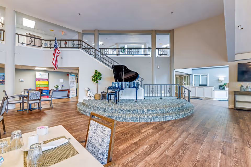 Spacious senior living facility common area with a grand piano on a raised circular platform, wooden flooring, dining tables and chairs, an American flag hanging near a staircase, and bright natural lighting.