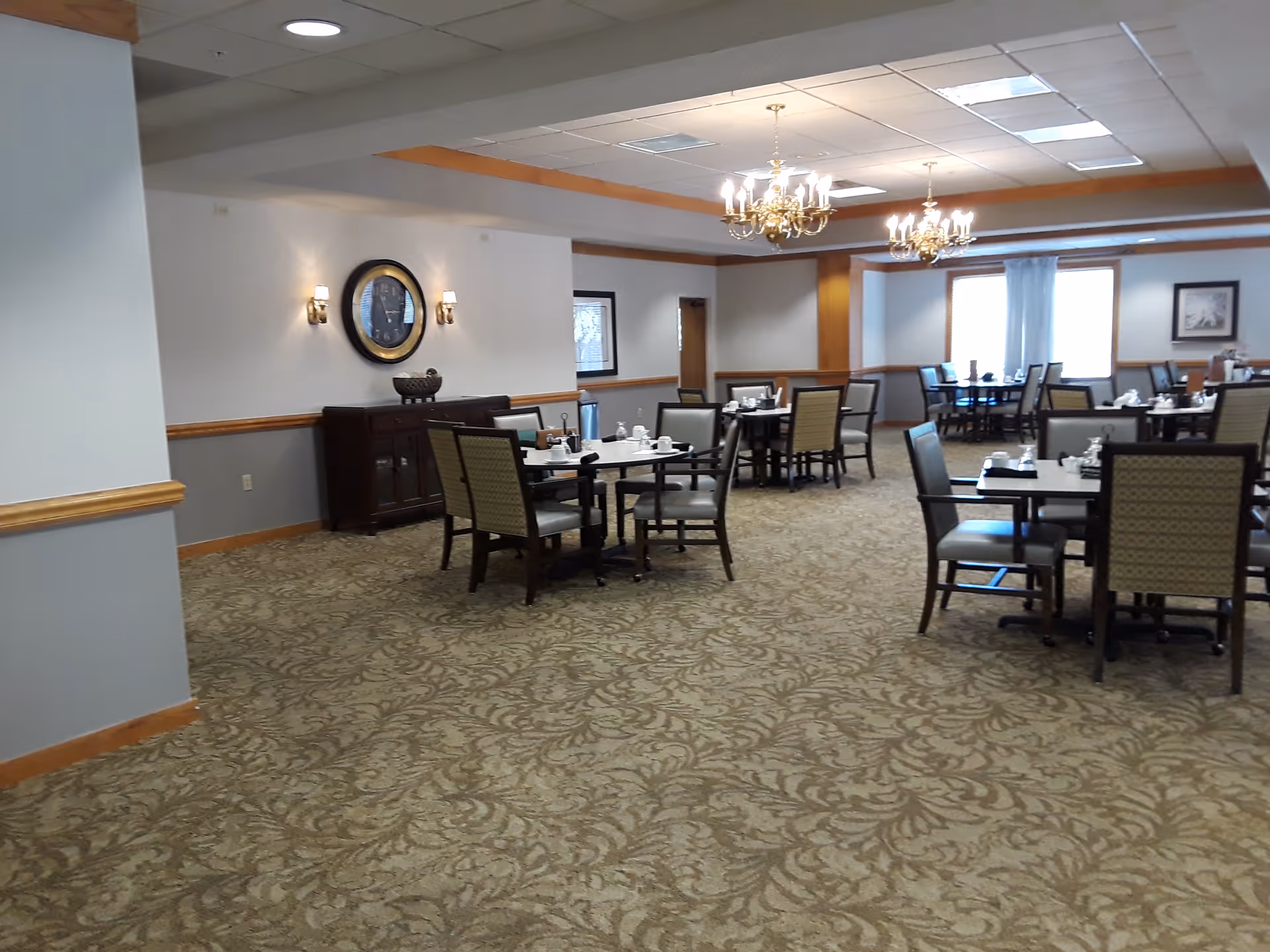 A spacious dining room with multiple tables and chairs arranged neatly. The room features patterned carpet flooring, white walls with wooden trim, two chandeliers hanging from the ceiling, a large clock on the wall, and windows with curtains allowing natural light in.
