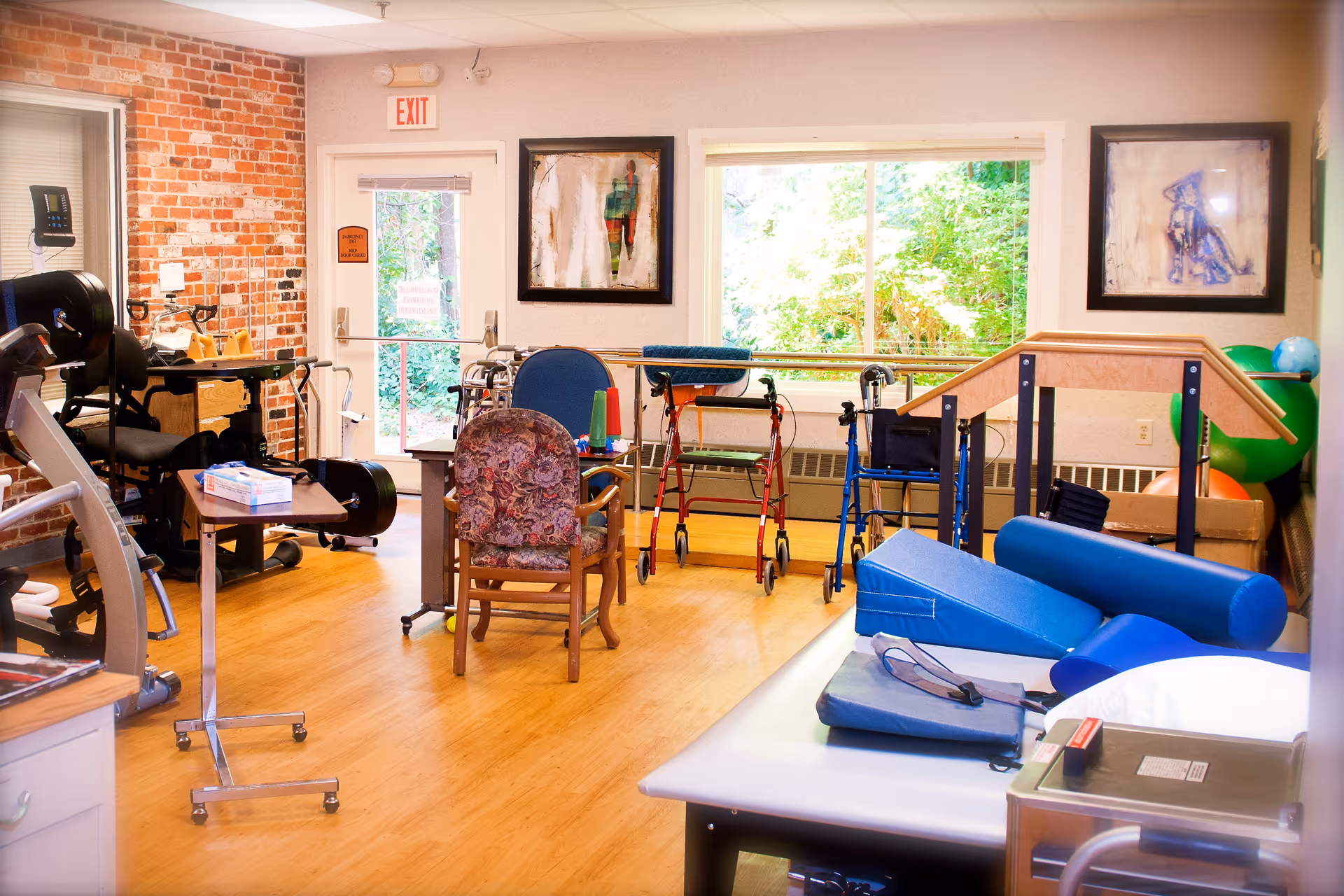 A bright therapy or rehabilitation room with exercise equipment, walkers, chairs, and therapy mats. The room has wooden flooring, a brick accent wall, large windows showing greenery outside, and framed artwork on the walls.