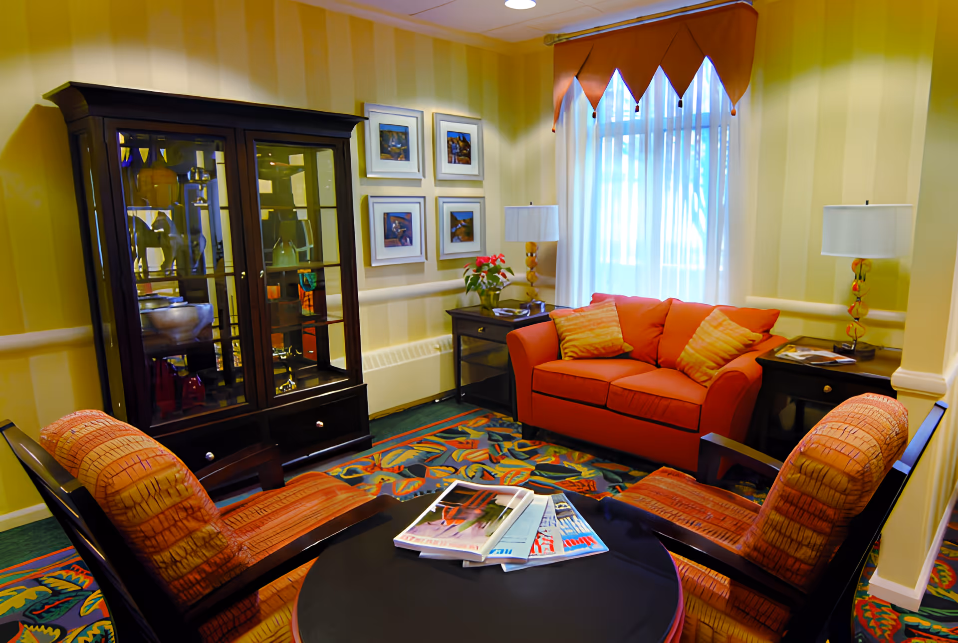 A cozy sitting area with an orange loveseat and two patterned armchairs around a round black coffee table with magazines on it. Behind the loveseat is a window with sheer white curtains and an orange valance. On either side of the loveseat are small dark wood side tables with lamps and a plant. A dark wood display cabinet with glass doors is against the wall, and four framed pictures hang on the wall above a white chair rail. The room has yellow striped wallpaper and a colorful patterned carpet.
