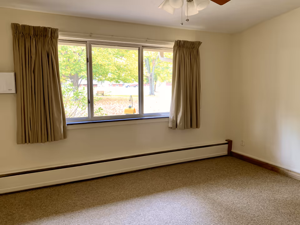 Empty room with beige carpet, cream-colored walls, a large window with beige curtains, and a ceiling fan with lights. Outside the window, green trees and a park bench are visible.