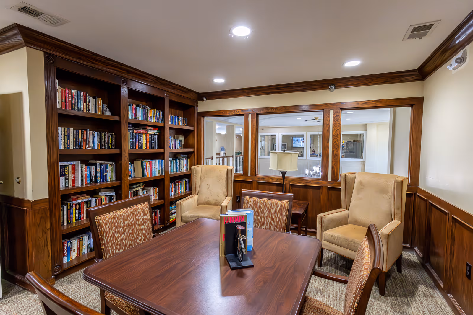 Wood-paneled communal reading room with built-in bookshelves, a central table and upholstered armchairs.
