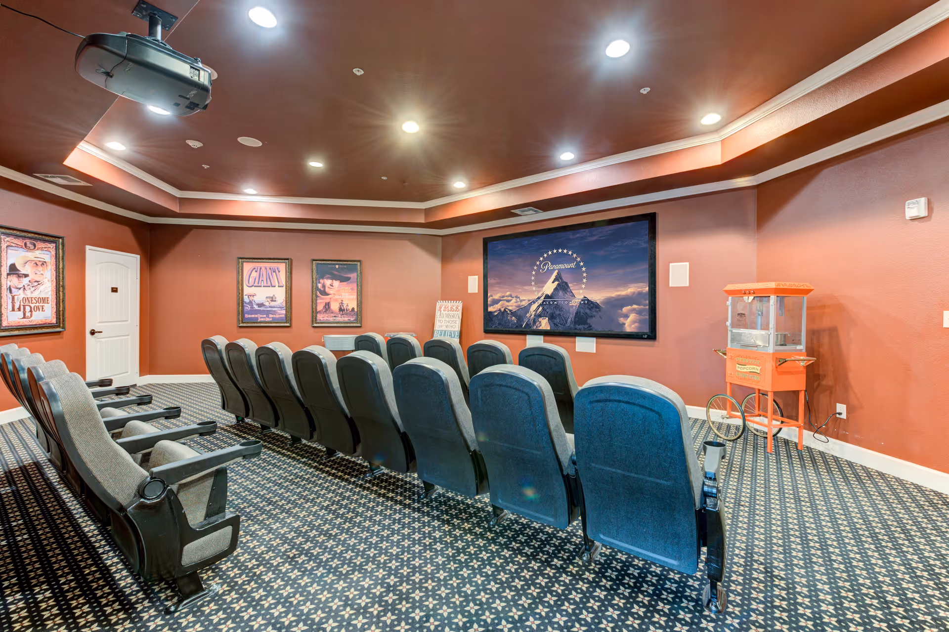 A cozy senior living facility movie theater room with rows of comfortable theater seats facing a large screen displaying the Paramount logo. The room has warm red walls, framed vintage movie posters, a popcorn machine on the right side, and recessed ceiling lights.