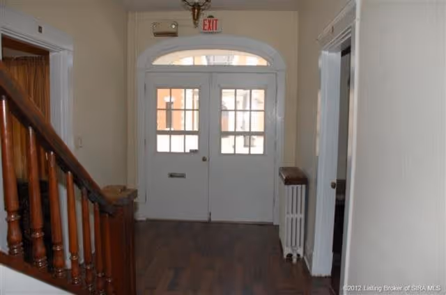 Interior view of a hallway with wooden flooring, a staircase with wooden banisters on the left, white double doors with glass panes at the end, and an exit sign above the doors. There is a radiator on the right side and doorways leading to other rooms on both sides of the hallway.