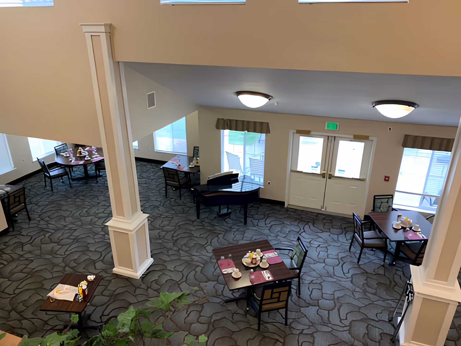 Interior view of a dining area in a senior living facility with several tables and chairs arranged on a patterned carpet floor. Each table is set with placemats, napkins, and condiments. There are large windows and double doors letting in natural light, and a piano is visible near the back wall.