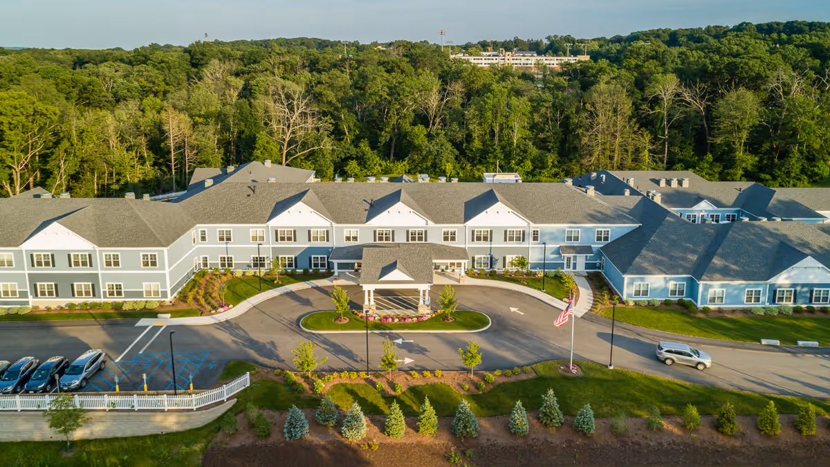 Aerial view of a large senior living facility named The Branches of Marlboro, featuring a two-story building with gray siding and white trim, surrounded by landscaped greenery and trees. There is a circular driveway with a covered entrance, several parked cars, and an American flag on a flagpole near the entrance.