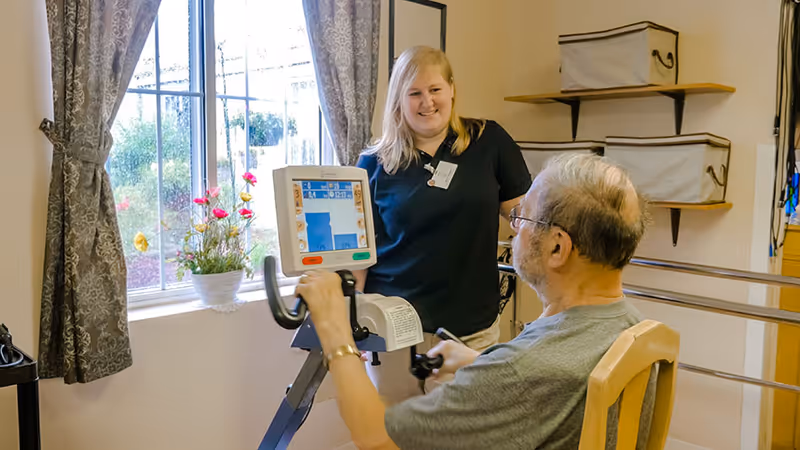 A man using a stationary exercise bike indoors while a smiling female caregiver or therapist watches and supports him. The room has a window with curtains, a flower pot on the windowsill, and shelves with storage bins on the wall.