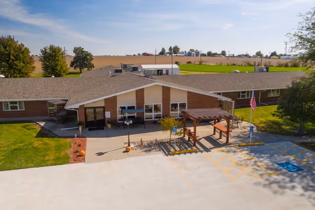 Exterior view of a single-story brick building with a gable roof, a small covered entrance, and a wooden pergola over a seating area. There are two handicap parking spaces in front, an American flag on a pole, and a rural landscape with fields and trees in the background under a clear sky.