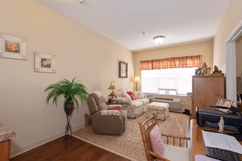 A cozy living room with beige walls and wooden flooring. The room features a floral patterned sofa with matching ottoman, a beige recliner with a lace doily on the armrest, a wooden side table with a lamp, and a wooden cabinet with framed photos on top. A large window with blinds and a red plaid valance lets in natural light. There is a patterned area rug covering part of the floor and a wooden desk with a chair, printer, and papers in the foreground.