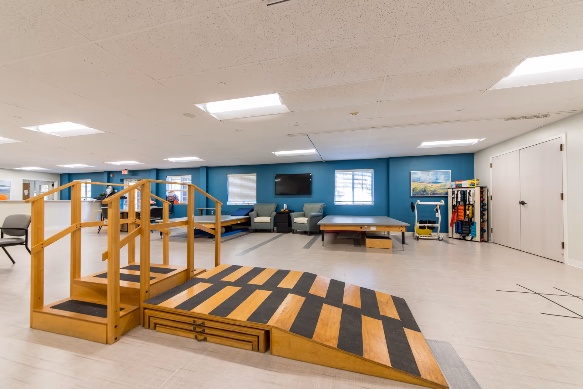 A spacious rehabilitation or physical therapy room with a wooden ramp and stairs in the foreground, two therapy tables, two armchairs, a TV mounted on a blue accent wall, and various exercise equipment and supplies neatly organized on shelves. The room has large windows allowing natural light and a light-colored floor.