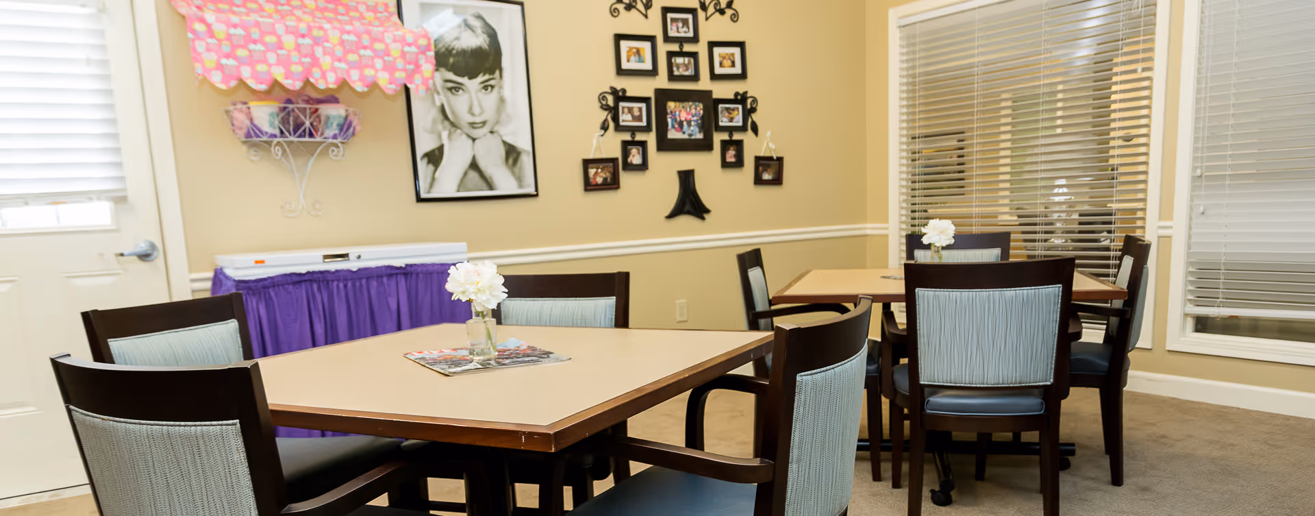 A cozy dining area with two square tables, each surrounded by four wooden chairs with light blue cushions. Each table has a small vase with white flowers and a magazine on it. The walls are beige with white trim, decorated with a black and white portrait of a woman and a collage of framed photos. There is a window with blinds and a door with a window blind partially closed. A purple skirted table with a pink cupcake-patterned cover is against the wall.
