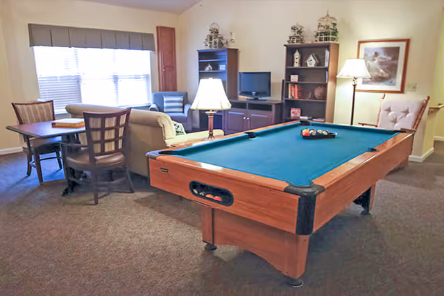Community lounge with a pool table in the foreground, seating area, TV, and a table by a bright window.