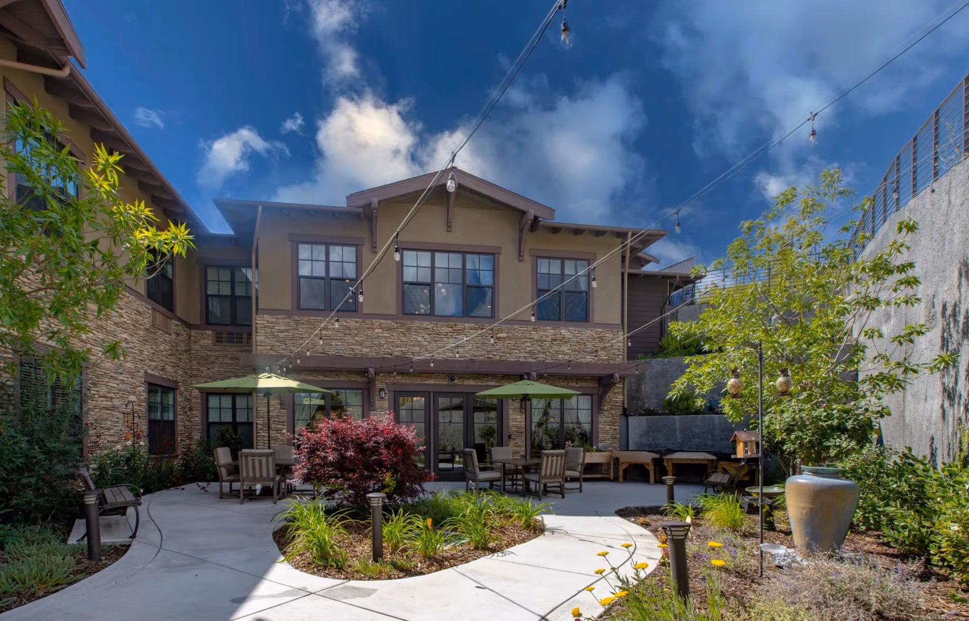 Outdoor courtyard area of a senior living facility with a paved walkway, garden beds with plants and flowers, patio tables with umbrellas, chairs, string lights overhead, and a two-story building with stone and beige exterior walls in the background under a partly cloudy sky.