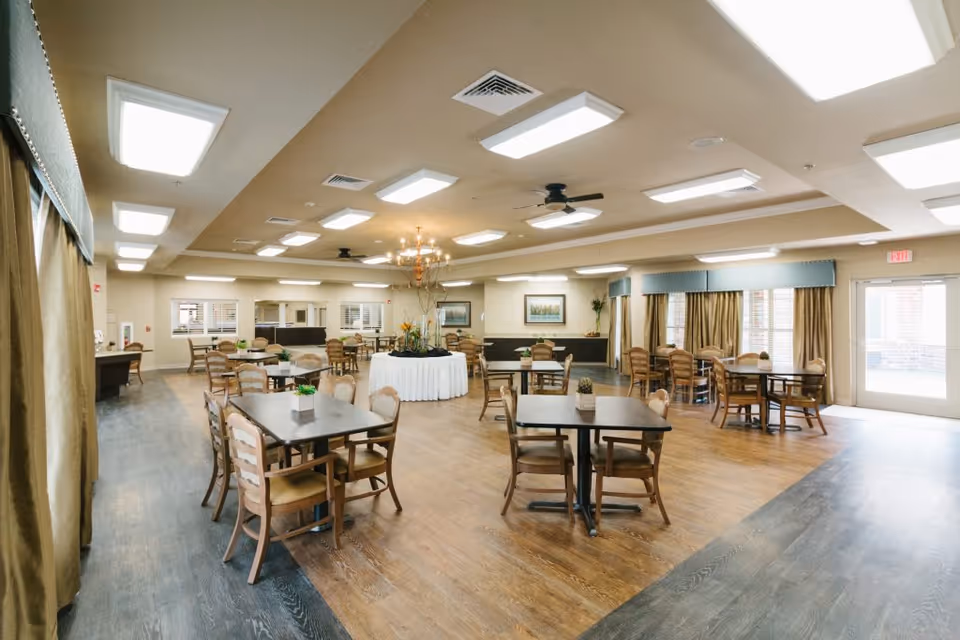 A spacious dining room with multiple wooden tables and chairs arranged neatly. The room has large windows with curtains, ceiling lights, a chandelier, and ceiling fans. There is a round table with a white tablecloth and a floral centerpiece in the middle of the room.