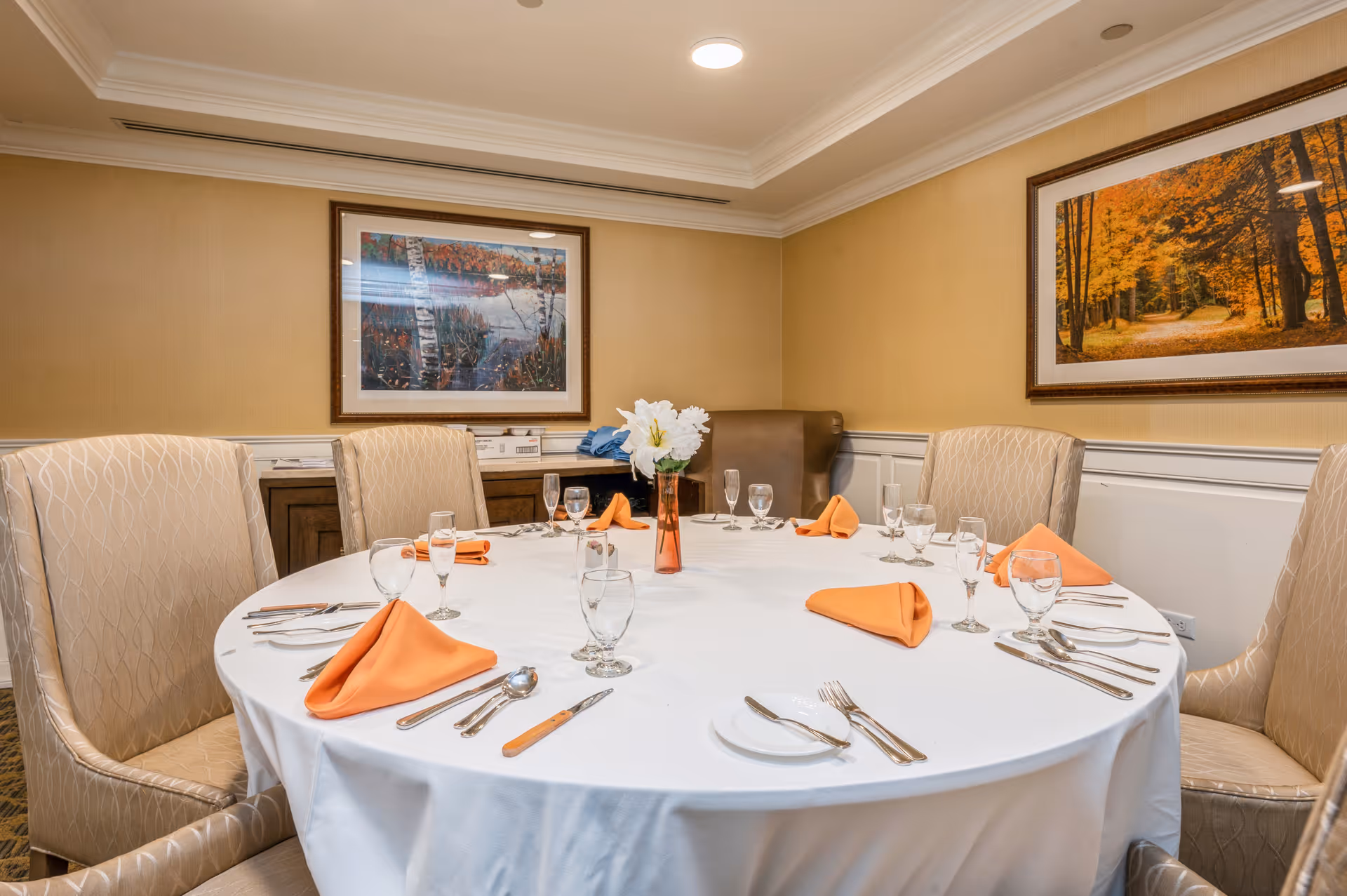 A round dining table set for six people with white tablecloth, orange folded napkins, glassware, and silverware in a warmly lit room. The room has beige walls with white wainscoting and two framed landscape paintings featuring autumn scenes. There are six upholstered chairs around the table and a small vase with white flowers in the center.