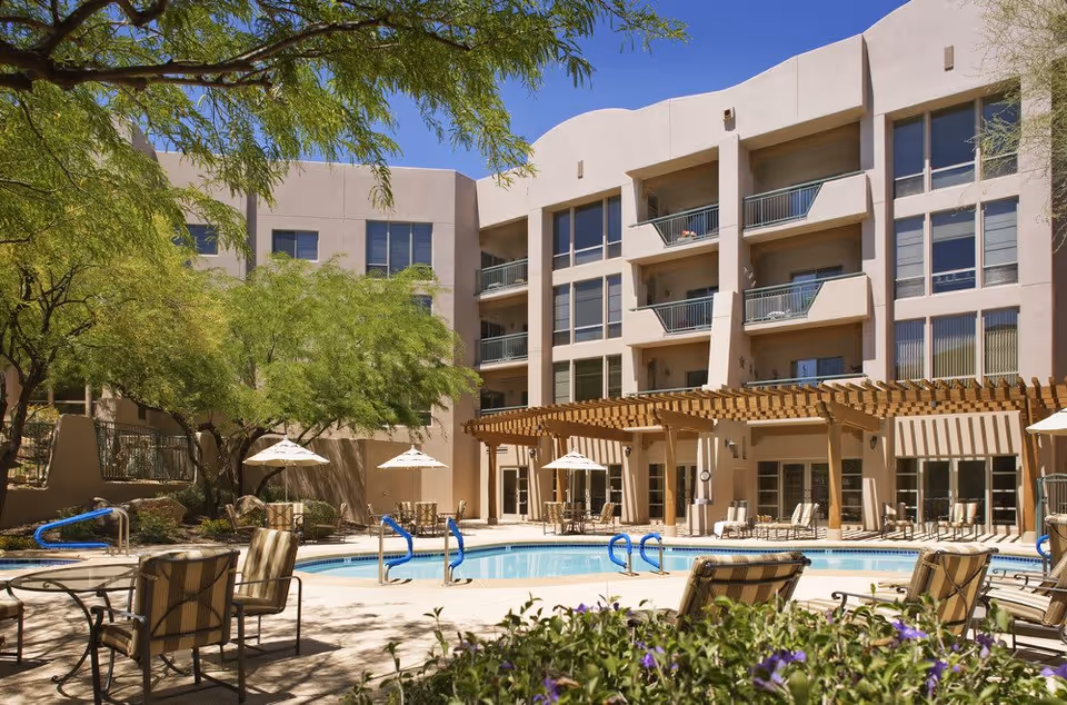 Outdoor view of a senior living facility courtyard with a swimming pool surrounded by lounge chairs and tables with umbrellas. The building has multiple floors with balconies and large windows, and there are trees providing shade around the pool area under a clear blue sky.