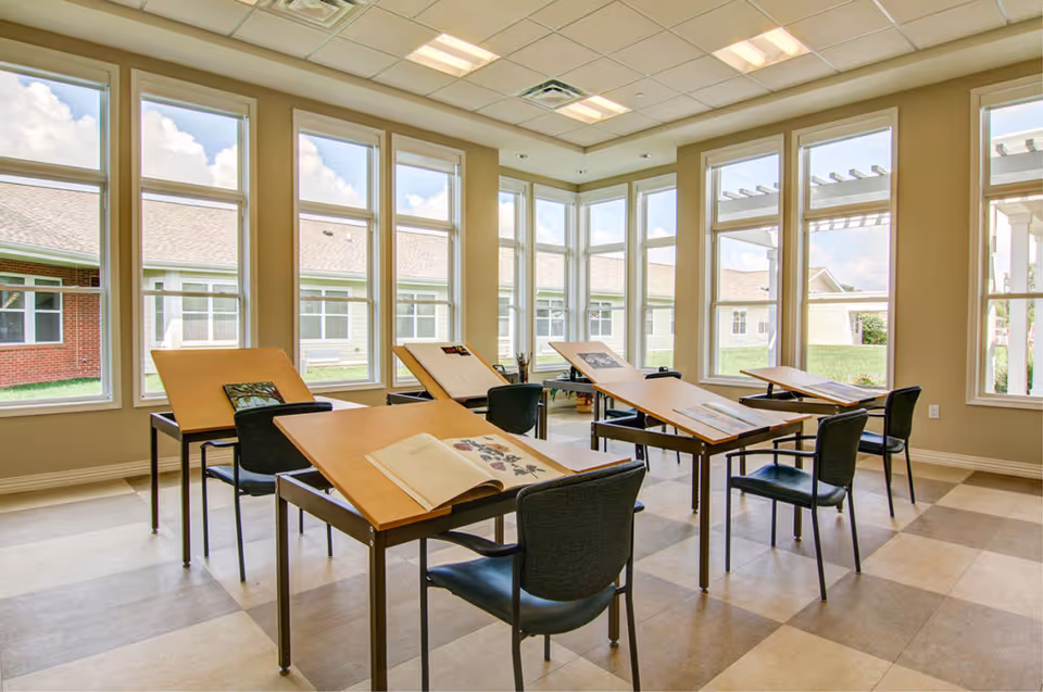 A bright room with large windows on two sides, featuring several drafting tables with angled surfaces and black chairs. The tables have open books and papers on them. Outside the windows, parts of a building and green grass are visible under a partly cloudy sky.