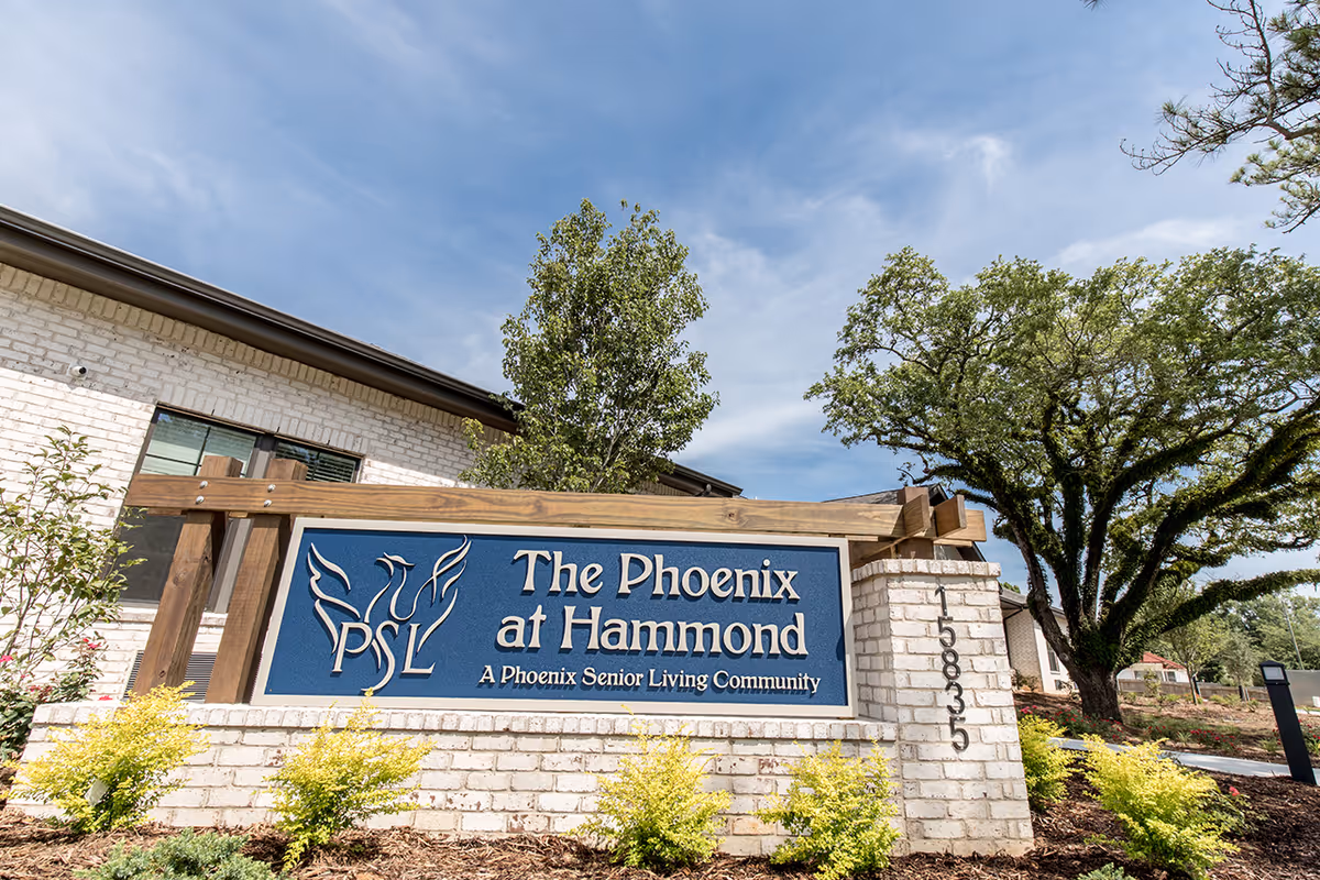 Outdoor view of the entrance sign for The Phoenix at Hammond, a senior living community, with a brick base and wooden frame surrounded by plants and trees under a partly cloudy sky.