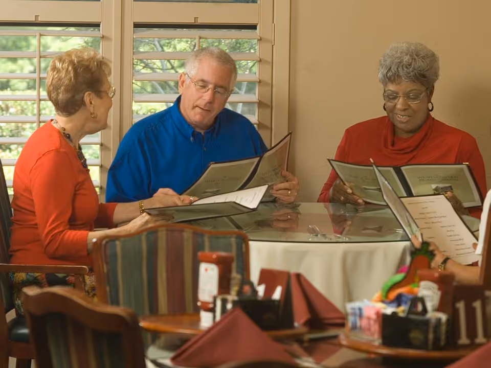 Three elderly people sitting around a round glass table in a dining area, looking at menus. The room has large windows with wooden shutters and the table is set with condiments and napkins.