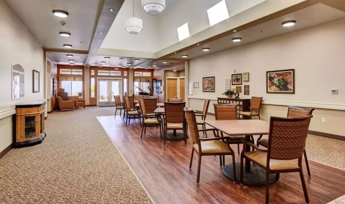 Spacious memory-care common area with multiple dining tables and chairs, seating alcoves, and skylights over a wood-look floor.